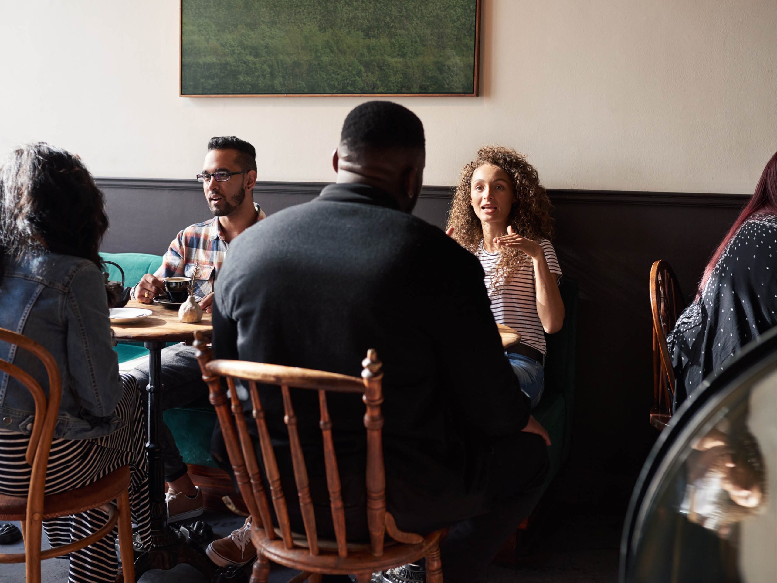 Flera personer i olika åldrar och med olika bakgrund sitter tillsammans vid olika bord på ett café. TIA Stockholm innebär att en rad olika organisationer arrangerar kostnadsfria språkcaféer för asylsökande ovh vissa massflyktingar i Stockholms län.