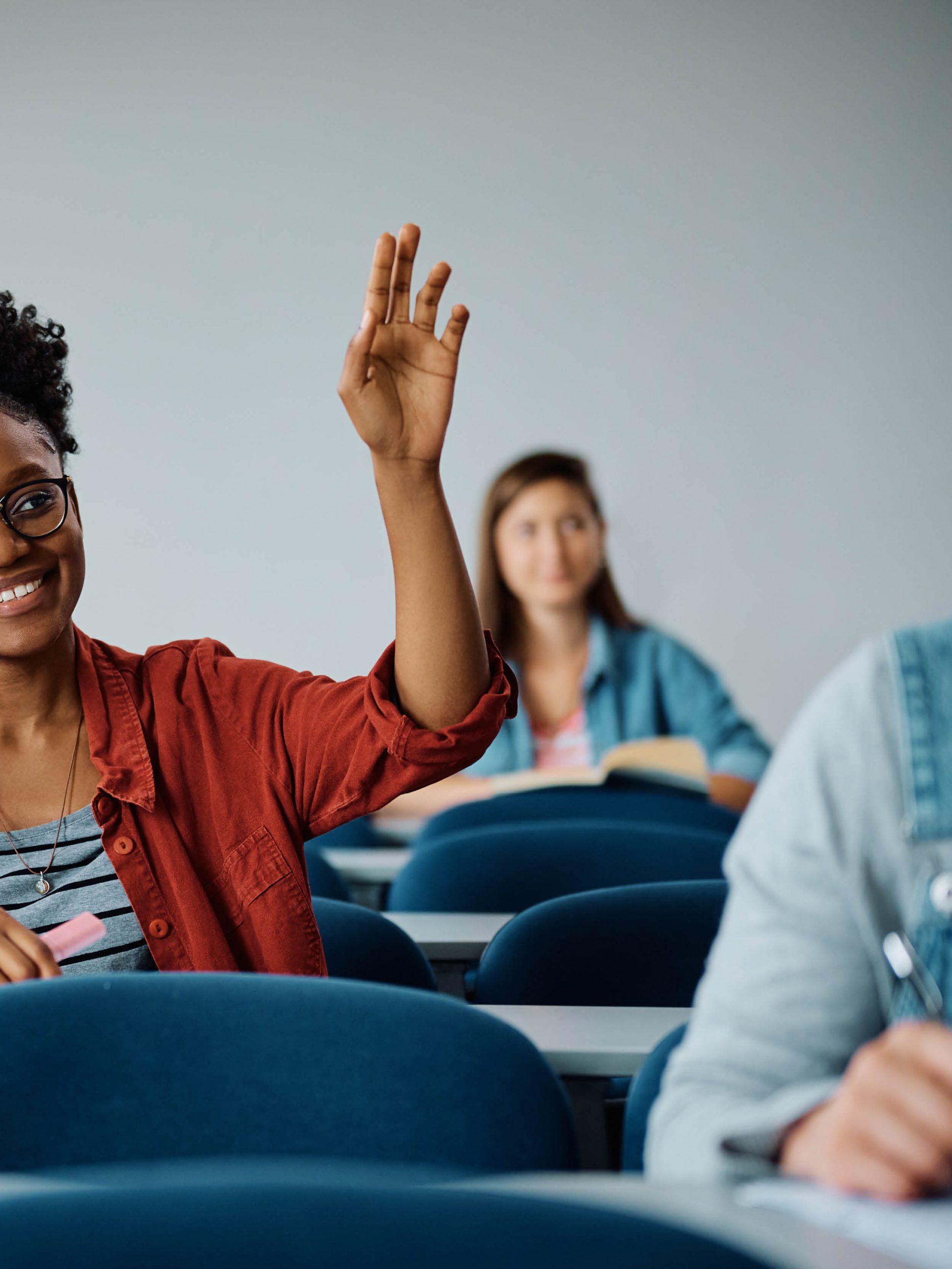 En kvinna i ett klassrum sitter centrerat i bilden med sin hand uppräckt under en lektion. I förgrunden och bakgrunden, oskärpa i bild, sitter andra personer på kursen och antecknar och lyssnar för att lära sig svenska.